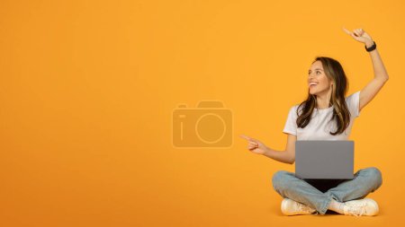 Téléchargez les photos : Joyeuse femme caucasienne en t-shirt blanc et jean bleu assis avec les jambes croisées sur le sol, à l'aide d'un ordinateur portable et gesticulant vers le haut avec une expression ravie sur un fond orange vif - en image libre de droit