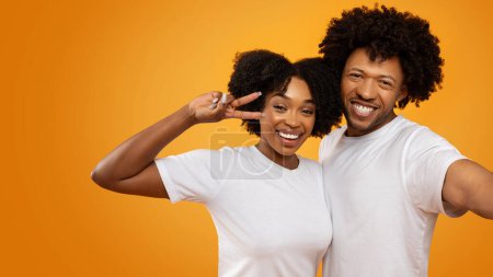 Téléchargez les photos : Joyeux jeune couple noir prenant selfie ensemble sur fond de studio orange. Joyeux joli millénium femme afro-américaine étreignant son mari et montrant geste de paix, panorama avec espace de copie - en image libre de droit