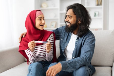 Téléchargez les photos : Heureux jeune couple musulman tenant test de grossesse positive et regardant les uns les autres tout en étant assis sur le canapé à la maison, mari arabe embrassant sa joyeuse épouse, Célébration en attente bébé, Espace libre - en image libre de droit