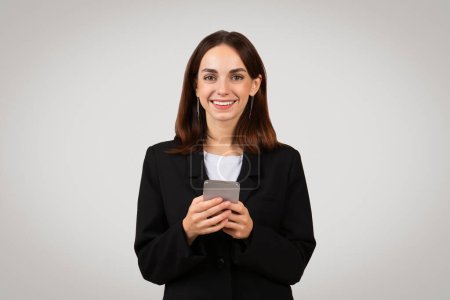 Photo for Cheerful millennial caucasian businesswoman in a sophisticated black suit engaging with her smartphone, possibly receiving good news or browsing through successful work results - Royalty Free Image