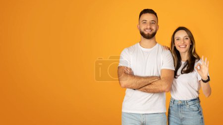Photo for Smiling confident caucasian man with crossed arms and positive woman making an OK sign, both in white t-shirts and jeans, expressing approval and satisfaction against an orange background - Royalty Free Image