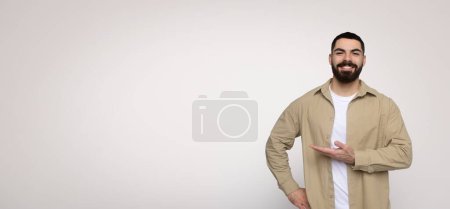Photo for Cheerful man with a beard presenting with his hand to the side, displaying an empty space for text or product, wearing a beige shirt and a white t-shirt on a wide background - Royalty Free Image