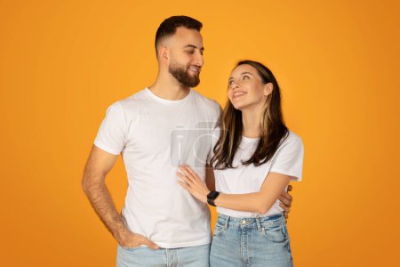Téléchargez les photos : Affectueux jeune couple caucasien debout près, avec la main des hommes sur sa hanche et la femme s'appuyant sur lui, souriant et se regardant sur un fond orange - en image libre de droit