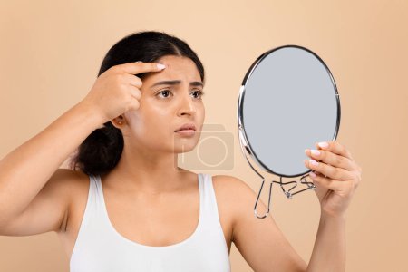 Photo for Concerned young indian woman closely inspecting her forehead in handheld mirror, worried female noticing pimple on face, suffering skin problems, standing against beige background, closeup - Royalty Free Image