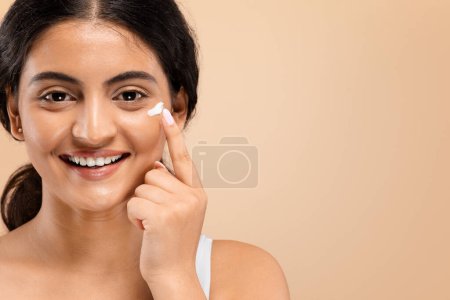Cheerful young indian woman applying dab of moisturizing cream on her face, happy beautiful eastern female promoting skincare routine with radiant smile, standing against beige background, copy space