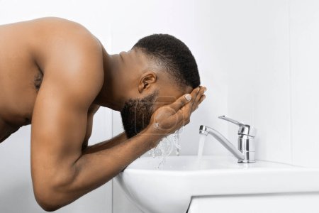 Téléchargez les photos : Jeune homme afro avec torse musculaire nu lavage visage dans l'évier à la salle de bain. Concept d'hygiène du matin, espace de copie - en image libre de droit