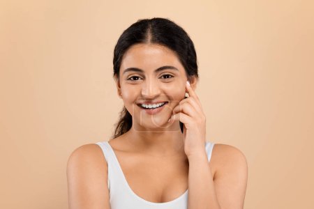 Foto de Mujer india alegre en camiseta blanca tocando su cara con una sonrisa suave, mujer oriental atractiva feliz de pie contra el fondo beige del estudio, personificando la belleza relajada y el autocuidado - Imagen libre de derechos