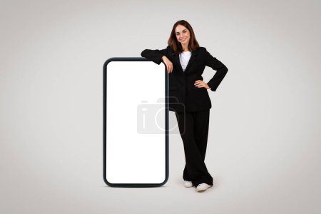 Photo for Confident millennial caucasian professional woman in a tailored suit leans on a giant smartphone with a blank display, illustrating the concept of tech-savvy business solutions - Royalty Free Image