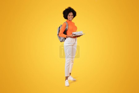 Photo for Confident young woman with a beautiful carrying a grey backpack and a stack of notebooks, dressed in an orange turtleneck and white pants, against a yellow background, studio - Royalty Free Image