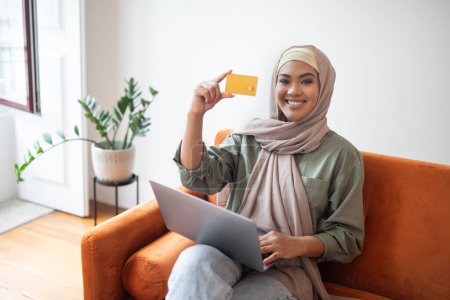 Photo for Digital Shopping. Happy Islamic woman in headscarf showing her credit card, makes online purchases via laptop in her living room, combining modern retail with casual lifestyle. Concept of ecommerce - Royalty Free Image