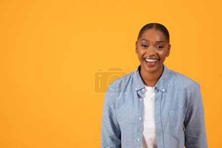 Téléchargez les photos : La beauté moderne. Femme noire joyeuse pose avec confiance en studio, mettant en valeur sa tenue décontractée en denim sur fond jaune, souriant à la caméra, exprimant bonheur et positivité. Espace libre - en image libre de droit