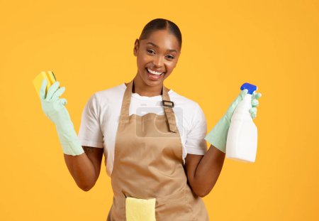 Photo for Spring cleanup. Cheerful African American millennial woman showcasing cleaning products in yellow studio setting, posing in apron and holding sponge and detergent, ready for house chores - Royalty Free Image