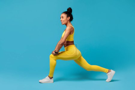 Photo for Active lifestyle, workout. Millennial black sportswoman in yellow fitness outfit lunging, flexing and strengthening legs, doing exercises in studio on blue backdrop. Sport motivation - Royalty Free Image