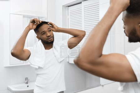 Téléchargez les photos : Millénaire afro homme peigner les cheveux et regarder dans le miroir dans la salle de bain. Homme dans la salle de bain concept - en image libre de droit