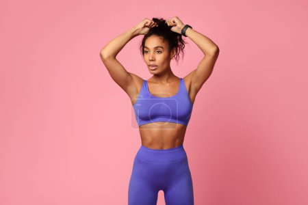 Photo for Fitness lifestyle. Motivated African American athletic lady in purple sporty outfit, touching hair making ponytail, preparing for gym workout, standing over pink studio background - Royalty Free Image
