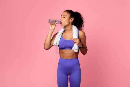 Téléchargez les photos : S'adapter jeune athlète femme noire en violet vêtements de sport eau potable, debout avec bouteille, rayonne de santé et de bien-être sur fond rose. Concept de fitness et d'hydratation saine pour la perte de poids - en image libre de droit
