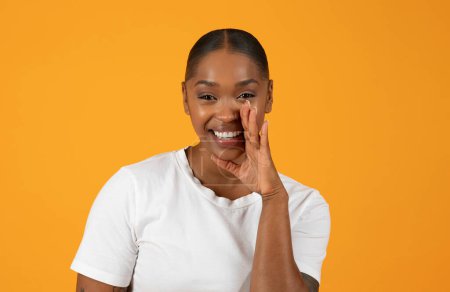 Téléchargez les photos : Femme afro-américaine joyeuse se tient sur fond orange, tenant sa main près de la bouche, partageant un secret ou une offre avec un sourire captivant. Concept de beaucoup et offre spéciale - en image libre de droit