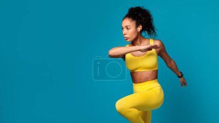 Photo for Cardio workout. Slim black lady with earbuds in yellow sporty outfit performing dynamic fitness exercise, jumping or running in studio over blue background. Panorama, empty space - Royalty Free Image