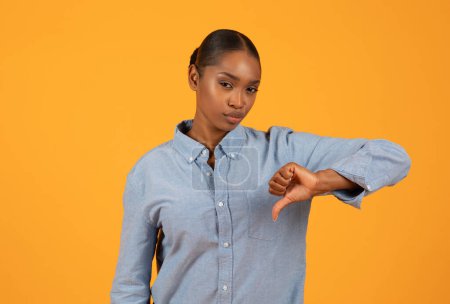 Téléchargez les photos : N'aime pas. Jeune femme noire exprime son mécontentement donnant pouces vers le bas geste en studio avec fond orange, véhiculant concept négatif ou désapprobateur dans la publicité moderne - en image libre de droit