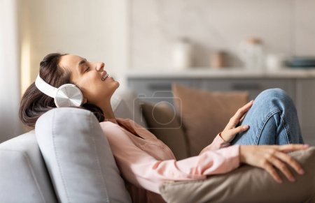 Téléchargez les photos : Femme satisfaite avec les yeux fermés savourant le moment, relaxante sur un canapé avec des écouteurs élégants, profondément immergée dans son répit rempli de musique - en image libre de droit