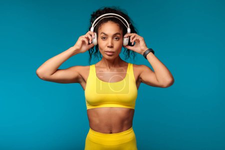 Photo for Fitness vibe. Motivated African American woman in yellow activewear stands confidently in studio, putting on wireless earphones, ready for healthy workout while listening to music on blue backdrop - Royalty Free Image