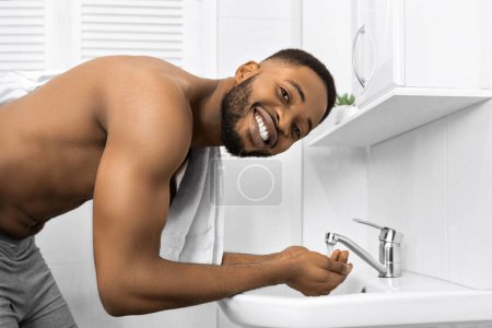 Photo for Smiling afro man with bare muscular torso washing face in bathroom, looking at camera Morning hygiene concept, copy space - Royalty Free Image