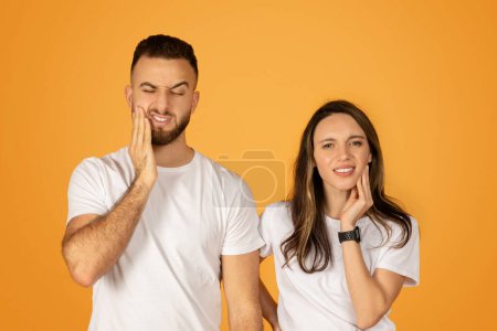 Foto de Joven hombre y mujer en camisas blancas mostrando dolor de muelas malestar, el hombre con una expresión dolorida sosteniendo su mejilla, y la mujer tocando suavemente la suya, sobre un fondo naranja - Imagen libre de derechos