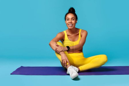 Photo for Workout vibes. Smiling black lady in yellow activewear with earbuds and tracker sits on mat, promoting fitness and leisure in studio over blue background. Copy space for gym ad - Royalty Free Image
