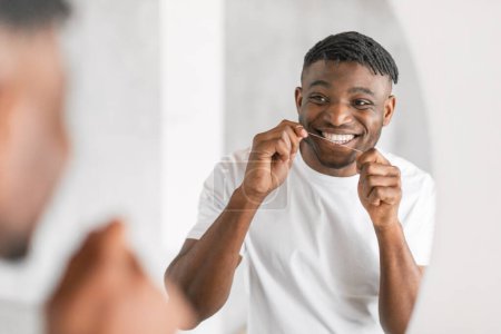 Lächelnder junger schwarzer Mann, der im Badezimmer mit Zahnseide vor dem Spiegel steht und sein Engagement für Mundpflege und Zahnhygiene betont, morgens Zähne mit Zahnseide putzt