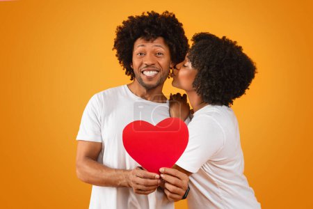 Photo for Romantic millennial african american couple celebrating anniversary on orange studio background. Loving black lady kissing her husband showing red paper heart. Love, relationships, marriage - Royalty Free Image