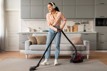 Photo for Contented woman wearing headphones and enjoys vacuuming tidy room with modern red vacuum cleaner, embodying joyful domestic chores - Royalty Free Image