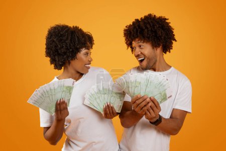 Photo for Cashback, lottery win, Emotional happy millennial african american spouses showing money euro banknotes and looking at each other, isolated on orange studio background - Royalty Free Image