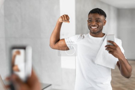 Téléchargez les photos : Sourire musclé noir guy prend selfie en utilisant son smartphone dans la salle de bain, montrant biceps regarder miroir réflexion, se préparer le matin et le partage choyer dans les médias sociaux - en image libre de droit