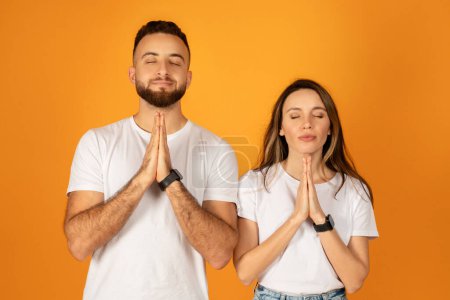 Photo for Peaceful caucasian young man and woman with eyes closed, hands pressed together in a prayer or meditative pose, dressed in white t-shirts, expressing calm, mindfulness against an orange background - Royalty Free Image