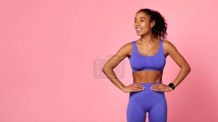 Téléchargez les photos : Modèle de fitness confiant. Femme afro-américaine athlétique en violet vêtements de sport se tient avec les mains sur les hanches, regarder de côté, mettant en valeur la force et le bien-être sur fond rose, panorama avec espace de copie - en image libre de droit