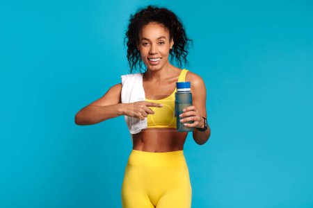 Photo for Fitness and hydration. Motivated sportswoman in blue studio setting points to a water bottle, radiating wellness and health, promoting exercise and fitness. Healthy lifestyle - Royalty Free Image
