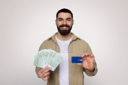 Téléchargez les photos : Un homme arabe du millénaire confiant avec une barbe en chemise beige détient des billets en euros dans une main et une carte bleue de crédit dans l'autre, représentant des options dans les transactions financières - en image libre de droit