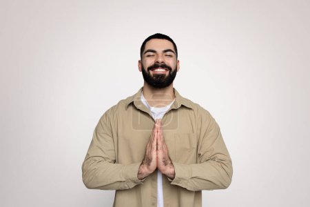 Téléchargez les photos : Un joyeux homme arabe millénaire avec une barbe soignée et un sourire éclatant fait un geste de prière dans une veste beige et un t-shirt blanc, exsudant de la positivité sur un fond propre, studio - en image libre de droit
