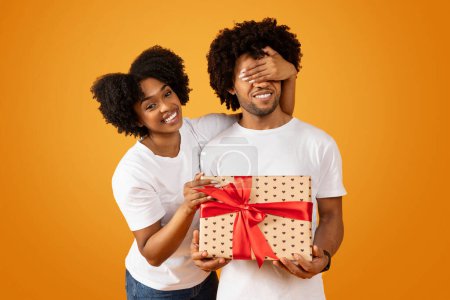 Téléchargez les photos : Aimer femme afro-américaine faire surprise d'anniversaire pour son mari, couvrant ses yeux et lui donnant cadeau boîte cadeau cadeau. Célébration anniversaire sur fond de studio orange - en image libre de droit