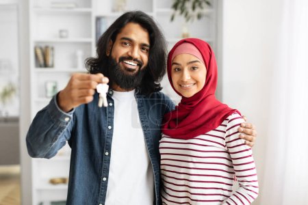 Foto de Nuevo concepto casero. Feliz pareja musulmana sosteniendo llaves de la casa, celebrando la compra de una nueva propiedad, felices cónyuges árabes propietarios de bienes de pie en el interior de la sala de estar, señora usando hijab, espacio libre - Imagen libre de derechos