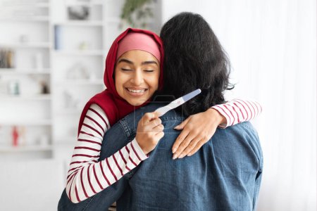 Happy Excited Young Muslim Woman Holding Positive Pregnancy Test And Hugging Husband, Joyful Islamic Family Embracing At Home, Celebrating Upcoming Parenthood, Closeup Shot With Copy Space