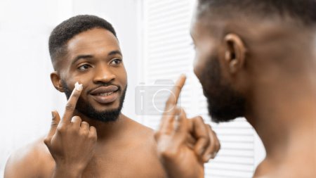 Homme afro souriant appliquant de la crème sur son visage dans la salle de bain, réfléchissant dans le miroir. Homme dans la salle de bain concept