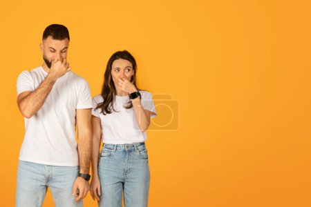Photo for Concerned young couple with hands over their mouths, displaying shock or surprise, wearing casual white t-shirts and blue jeans against an orange background with copy space - Royalty Free Image