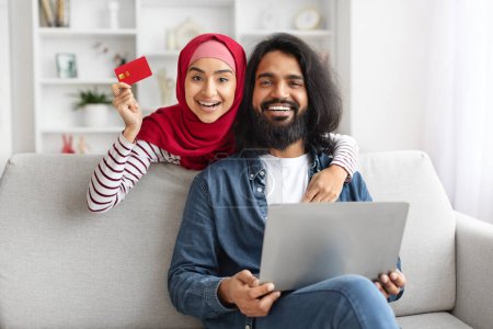 Photo for Online Shopping Concept. Happy Muslim Couple Using Laptop And Credit Card At Home, Cheerful Young Arabic Spouses Purchasing In Internet While Relaxing With Computer In Living Room, Copy Space - Royalty Free Image