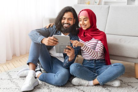 Téléchargez les photos : Heureux jeune couple musulman en utilisant une tablette numérique tout en se relaxant dans le salon ensemble, Souriant Arabe Conjoints Shopping en ligne, Naviguer sur Internet ou vérifier une nouvelle application, Gros plan - en image libre de droit
