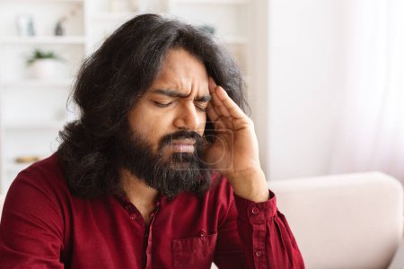 Photo for Closeup of unhealthy indian millennial man suffering from migraine, sitting with closed eyes on couch at home, touching his head, have headache, copy space. Healthcare, meteosensitivity - Royalty Free Image