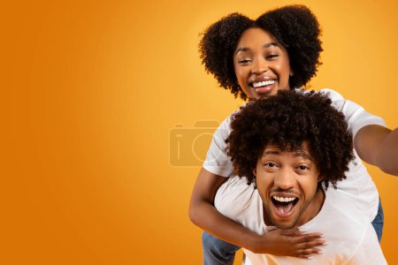 Photo for Cool millennial african american couple taking selfie on orange studio background. Cute pretty black lady piggybacking her boyfriend and holding camera, blank copy space - Royalty Free Image