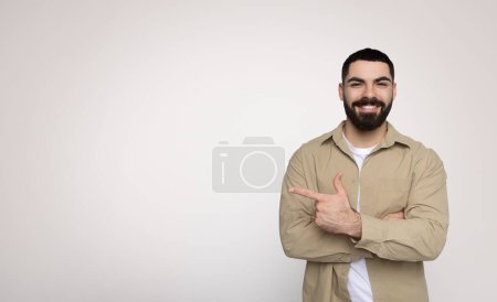 Téléchargez les photos : Joyeux et confiant homme barbu millénaire donnant un geste pouce vers le haut, vêtu avec désinvolture d'une chemise beige à manches longues sur un t-shirt blanc, sur un fond gris clair - en image libre de droit