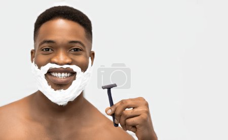 Photo for Smiling afro man with naked torso and shaving foam on his beard, holding razor, looking in mirror at bathroom. Comfort shaving concept, copy space - Royalty Free Image