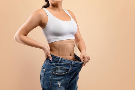 Photo for Fit woman confidently demonstrating her successful weight loss by pulling out oversized jeans, unrecognizable lady highlighting her slim figure, standing against soft beige studio background, copy - Royalty Free Image